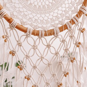 Puede incluir: Un atrapasue&ntilde;os de macram&eacute; blanco con un aro de madera y plumas blancas. El atrapasue&ntilde;os tiene un delicado dise&ntilde;o de crochet y est&aacute; decorado con peque&ntilde;as cuentas de madera.