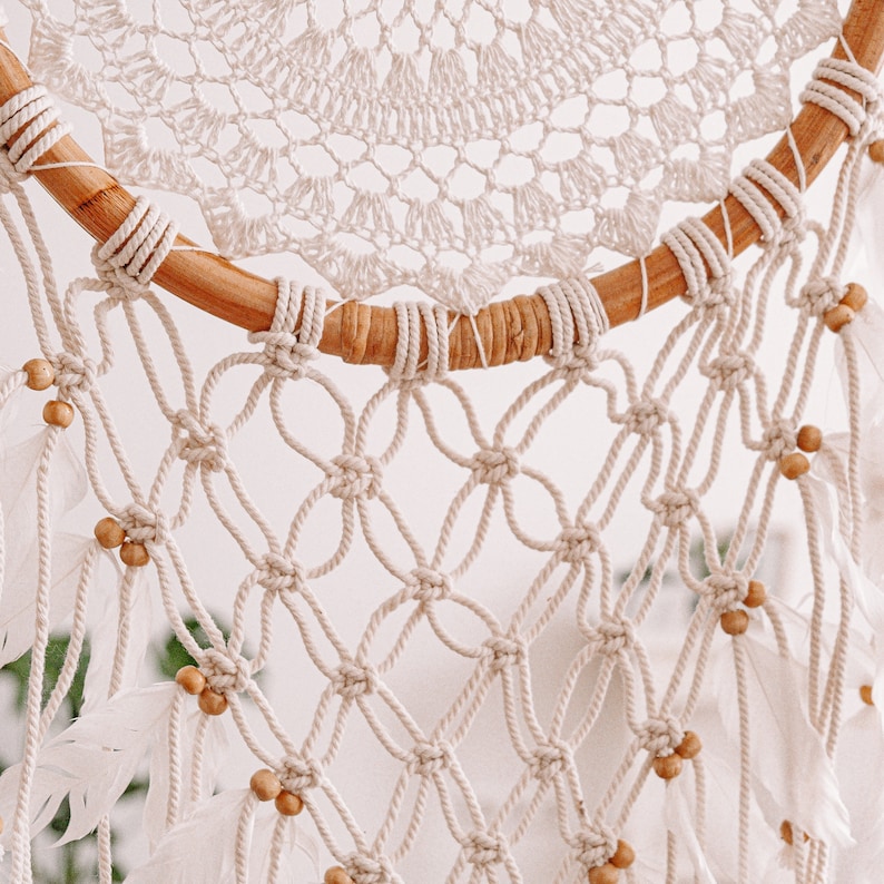 Puede incluir: Un atrapasue&ntilde;os de macram&eacute; blanco con un aro de madera y plumas blancas. El atrapasue&ntilde;os tiene un delicado dise&ntilde;o de crochet y est&aacute; decorado con peque&ntilde;as cuentas de madera.