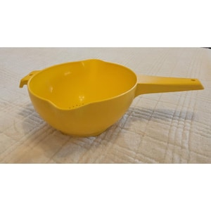 May include: A bright yellow plastic colander featuring a long, flat handle on one side and a smaller tab handle on the opposite side. The bowl has a rounded shape with a series of small drainage holes at the bottom and a smooth, matte finish.