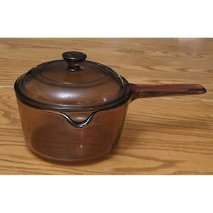 May include: A transparent amber-coloured glass saucepan with a matching lid and a long side handle. The pot features a rounded body with a small pouring spout on one side and a circular knob on the centre of the lid for easy lifting.