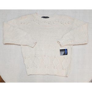 Könnte beinhalten: Ein cremefarbener Langarm-Strickpullover mit Rundhalsausschnitt sowie gerippten Bündchen und Saum. Das Kleidungsstück zeigt ein strukturiertes Muster mit erhabenen Bommel-Akzenten im Brust- und unteren Bereich sowie ein Rauten-Zopfstrickmuster.