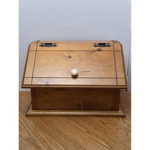 Può includere: Un portapane in legno marrone chiaro con coperchio frontale inclinato, un pomello rotondo bianco e due cerniere in metallo nero sulla parte superiore. La scatola presenta un design a cornice rettangolare inciso sul pannello frontale.