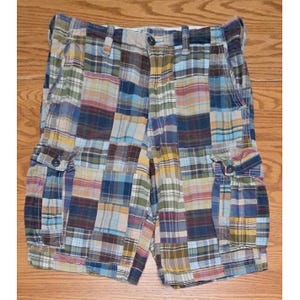 May include: Cargo shorts featuring a patchwork design made of various plaid fabric squares in shades of blue, brown, yellow, and white. The shorts include a button closure, a zipper fly, belt loops, and two side cargo pockets with button flaps.