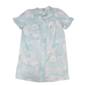 May include: A light blue housecoat featuring a white floral pattern, short sleeves, and a button-down front. The garment includes a round neckline with decorative white eyelet lace trim and two front patch pockets on the lower section.