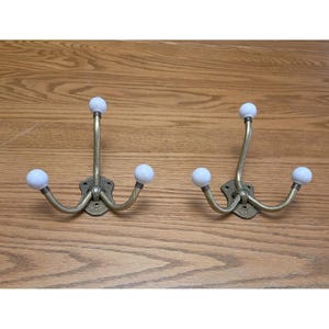 May include: Two wall-mounted coat hooks made of antique brass-toned metal. Each hook features a central vertical arm and two curved side arms, all topped with smooth white spherical knobs. The base of each hook has a decorative plate.