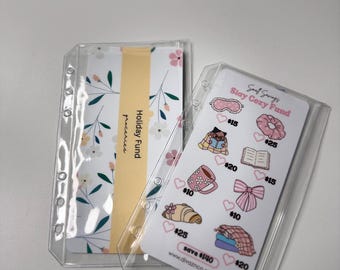 May include: Clear plastic binder inserts with printed cards. One insert has a floral design and the words "Holiday Fund groceries." The other insert has a "Stay Cozy Fund" card with illustrations and amounts. Website address is visible.