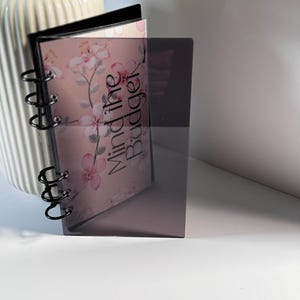 May include: A clear, dark gray planner with a black spiral binding. The planner is open, revealing pages with a floral design and the words "Mind the Budget" in a script font. The planner is standing upright.