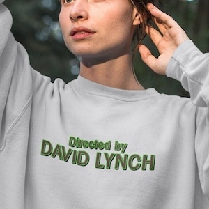 Puede incluir: Una sudadera blanca con texto verde que dice "Directed by DAVID LYNCH". La persona que lleva la sudadera está de pie en un bosque.