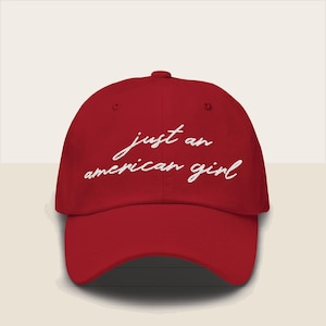 Puede incluir: Gorra de béisbol roja con visera curva. La gorra presenta el texto blanco en cursiva "just an american girl" bordado en la parte delantera. El sombrero es de color liso y tiene una parte superior redondeada.