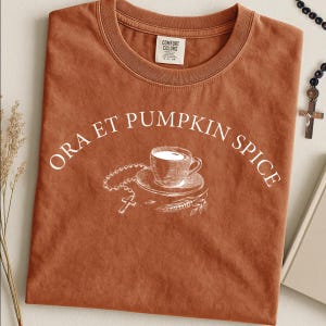 Può includere: Una t-shirt a maniche corte color ruggine con una stampa grafica bianca raffigurante una tazza di caffè, un rosario e spighe di grano. Il testo "ORA ET PUMPKIN SPICE" è stampato in un carattere bianco ad arco sopra la grafica.