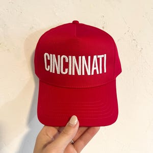 Könnte beinhalten: Eine rote Baseballkappe mit einem weißen, gestickten "CINCINNATI"-Logo.