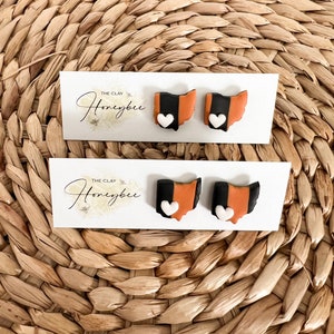 May include: A pair of orange, black, and white Ohio state-shaped earrings with a white heart on each. The earrings are on a white card with the text "The Clay Honeybee".