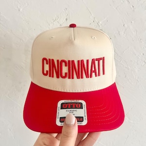 Puede incluir: Una gorra de béisbol beige y roja con la palabra "CINCINNATI" bordada en rojo en la parte delantera. La gorra tiene una visera roja y una etiqueta en blanco y negro que dice "OTTO" con las palabras "PREMIUM FABRIC", "STYLISH LOOK", "CLASSIC FIT", "MOST POPULAR FIT", "MILE", "MA" y "FORT" impresas en ella.