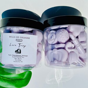May include: Two clear glass jars filled with lavender-colored wax melts. The jars have black lids and are labeled "WILLS DE COLOGNE" and "Love Trap". The wax melts are shaped like hearts, flowers, and other decorative shapes.