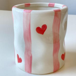 May include: A white ceramic vase with pink vertical stripes and red heart accents. The vase has a slightly irregular shape and a smooth, glossy finish. The design is simple and charming, suitable for home decor.