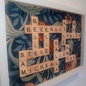 May include: A framed artwork with wooden Scrabble tiles spelling out names against a floral background. Names include BEVERLEY, LESLEY, STEVE, SAM, MICHAEL, LIAM, and YAZ. The frame is white.