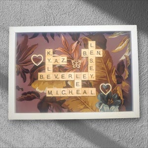 May include: A framed artwork with a floral design in brown, yellow, and purple. Wooden letter tiles spell out names, with two wooden heart accents. The frame is white.