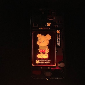 Puede incluir: Primer plano de una funda de teléfono con un diseño de oso de dibujos animados. El oso lleva un atuendo rojo y blanco y tiene una X cosida en los ojos. El texto "(ORIGINAL FAKE) COMPANION" está impreso en la funda.