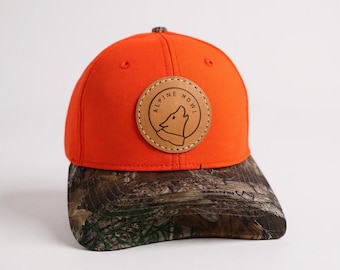 Gorra de camionero color naranja brillante con borde RealTree, cierre ajustable con parche de cuero y diseño de aullido alpino.