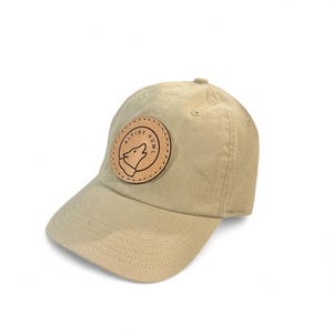 May include: A tan baseball cap with a curved brim. A circular leather patch on the front features a howling wolf graphic and the text "ALPINE HOWL". The hat is made of a durable fabric and has a classic, casual style.