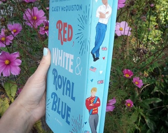 Può includere: Un libro rilegato in blu con il titolo "Red, White & Royal Blue" di Casey McQuiston. La copertina del libro presenta un design di fuochi d'artificio rosso, bianco e blu e due uomini in illustrazioni sul dorso.