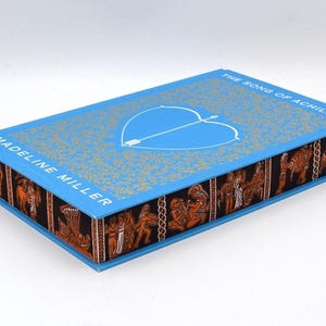 Könnte beinhalten: Ein blaues Hardcover-Buch mit dem Titel "Der Gesang des Achilles" von Madeline Miller. Der Umschlag zeigt ein weißes Herz mit einem Pfeil hindurch und einen gold-schwarzen Musterrahmen.