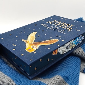May include: A hardback book titled "Harry Potter and the Philosopher's Stone" with a navy blue cover, gold lettering, and a golden owl illustration. The book is on a blue and grey striped scarf.