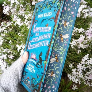 Könnte beinhalten: Ein Hardcover-Buch mit dem Titel "Kompendium der Verlorenen Geschichten" von Emily Wildes und Heather Fawcett. Das Buch hat einen türkisfarbenen Einband mit Illustrationen einer schwarzen Katze, Bienen und floralen Elementen. Das Buch wird vor einem Hintergrund aus weißen Blumen gehalten.