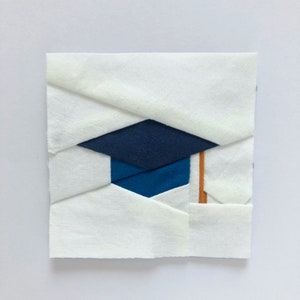 Op de afbeelding: Een wit vierkant van stof met een blauw en donkerblauw afstudeerhoedontwerp. De hoed heeft een bruine kwast.