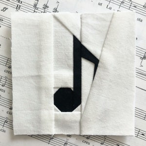 May include: A white fabric square with a black musical note design. The note is made of fabric pieces sewn together.