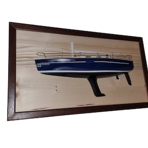 Könnte beinhalten: Ein Wandbild in einem Holzrahmen mit einem blauen und weißen Segelbootmodell auf einem braunen Holzsockel. Das Segelboot hat eine weiße Kabine und ein braunes Geländer. Das Modell ist auf einer Holzplatte mit einem braunen Rahmen montiert.