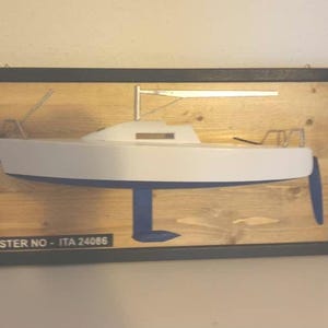 Könnte beinhalten: Ein weißes Segelbootmodell, das auf einer Holzplatte mit schwarzem Rahmen montiert ist. Die Platte trägt den Text "MISTER NO - ITA 24086".