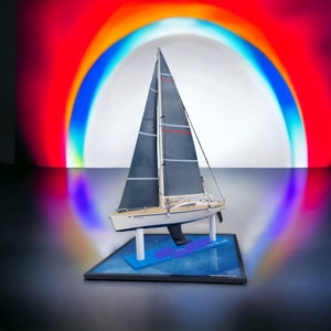 May include: A model sailboat with a black and gray sail, a white hull, and a blue base. The sailboat is displayed on a black base with a blue water-like surface. The sailboat is in front of a rainbow-colored background.