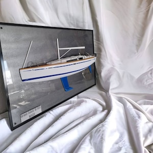 Könnte beinhalten: Ein weißes Segelbootmodell mit blauen Akzenten wird in einem schwarzen Rahmen auf grauem Hintergrund präsentiert. Das Modell ist mit "Grand Soleil 40 1982 MAGIA" beschriftet.