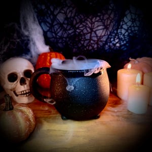 May include: A black cauldron-shaped mug with a textured finish, featuring a single handle and three small feet. The mug is filled with swirling white vapor, surrounded by a human skull, orange pumpkins, and two lit white pillar candles.