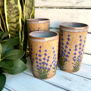 May include: Three ceramic mugs with a speckled brown glaze and a lavender floral design. The mugs are different sizes and are arranged on a white wooden surface.