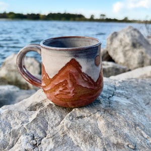 May include: A ceramic mug with a rounded body and a curved handle. The exterior features a two-tone glaze with a textured, reddish-brown mountain range design at the base and a smooth, light gray finish on the upper portion and rim.
