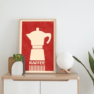 Pu&ograve; includere: Una stampa incorniciata con uno sfondo rosso e una silhouette bianca di una caffettiera. Il testo "Kaffee" &egrave; stampato in bianco sotto la caffettiera.