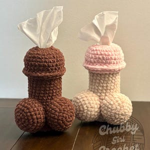 May include: Two handmade crochet tissue box covers shaped like male anatomy. One is brown, the other is beige with a pink tip. Each has a white tissue emerging from the top. The text "Chubby Girl Crochet" is visible.