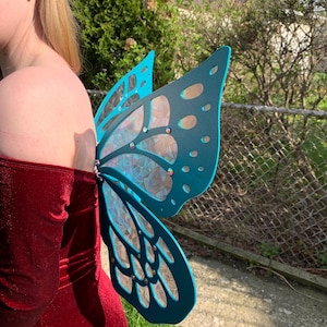 May include: A pair of teal butterfly wings featuring a cut-out frame design with iridescent, stained-glass style panels. The wings are adorned with small, reflective circular accents embedded within the structure, creating a shimmering effect.