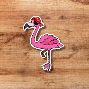 Puede incluir: Una pegatina de flamenco rosa con temática pirata. El flamenco lleva un pañuelo rojo y un parche en el ojo. La pegatina está sobre una superficie de madera.