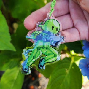 May include: A green and blue acrylic keychain featuring a cartoon character with a weapon. The character is green with black eyes and is holding a blue gun. The keychain has a green clasp and a green string.