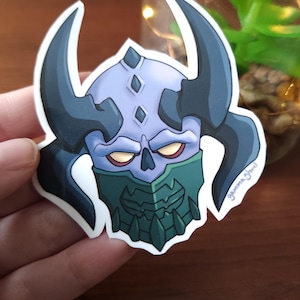 May include: A sticker featuring a stylized illustration of a horned skull with a blue and green color scheme. The skull has large black horns, yellow eyes, and a detailed green mask. The sticker is held in a hand.