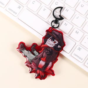 Peut inclure: Un porte-clés en acrylique rouge représentant un personnage de dessin animé portant un chapeau et un manteau. Le personnage a les yeux rouges et est allongé. Le porte-clés a un fermoir en métal noir et repose sur un clavier blanc.