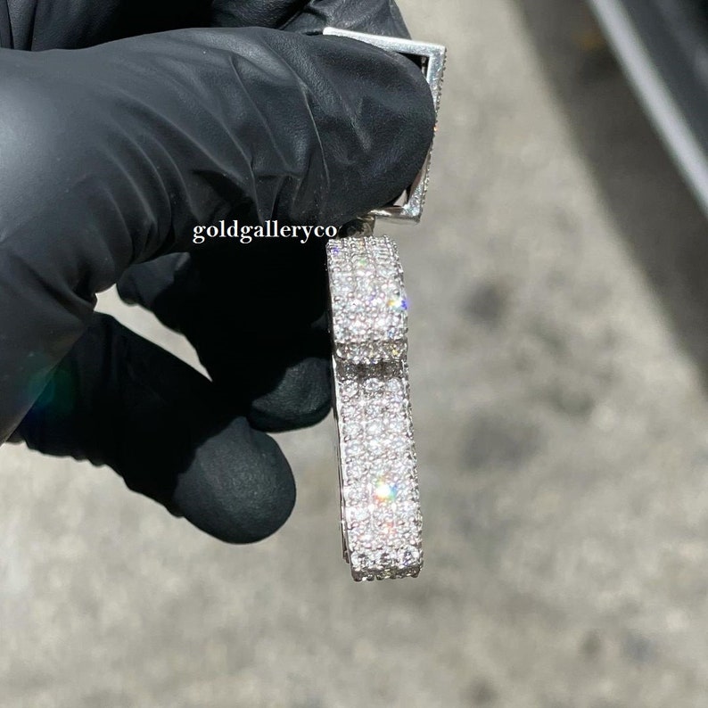May include: A silver pendant with a rectangular shape, covered in small, clear diamonds. The pendant is held in a gloved hand.