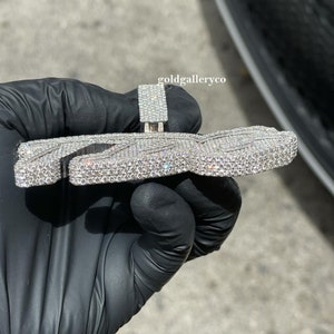 May include: A silver pendant shaped like the letter "Z" with a diamond-encrusted design. The pendant is held in a gloved hand.