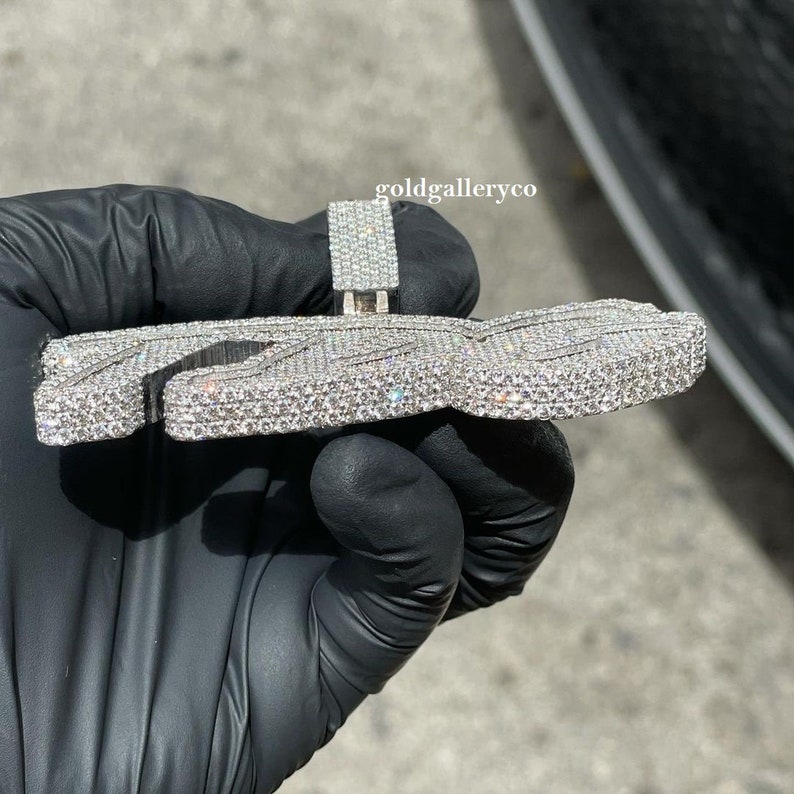 May include: A silver pendant shaped like the letter "Z" with a diamond-encrusted design. The pendant is held in a gloved hand.