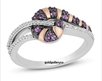 May include: A silver ring with a black and rose gold swirl design. The swirl is set with purple gemstones and the band is accented with small white gemstones. The ring is engraved with the text "goldgalleryco".