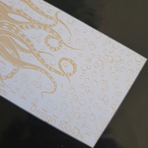 Puede incluir: Una tarjeta blanca con un diseño en relieve dorado de un pulpo con tentáculos y burbujas. El texto "Design 100434" está impreso en dorado en la parte inferior de la tarjeta.