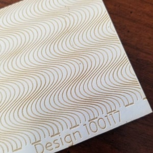 Op de afbeelding: Een wit vel papier met een geëmbost golvend patroon op het oppervlak. De tekst "Design 100117" is gedrukt in de rechterbenedenhoek.